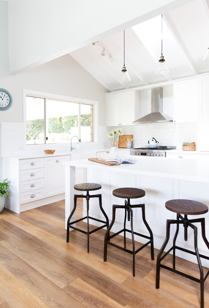 This is an example of a country l-shaped eat-in kitchen in Gold Coast - Tweed with a farmhouse sink, shaker cabinets, white cabinets, quartz benchtops, white splashback, subway tile splashback, stainless steel appliances, vinyl floors, with island and brown floor.