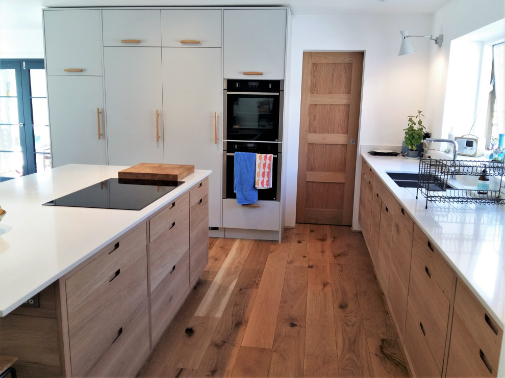 Photo of a large contemporary l-shaped open plan kitchen in Cornwall with a double-bowl sink, flat-panel cabinets, medium wood cabinets, solid surface benchtops, black appliances, medium hardwood floors, a peninsula, brown floor and white benchtop.
