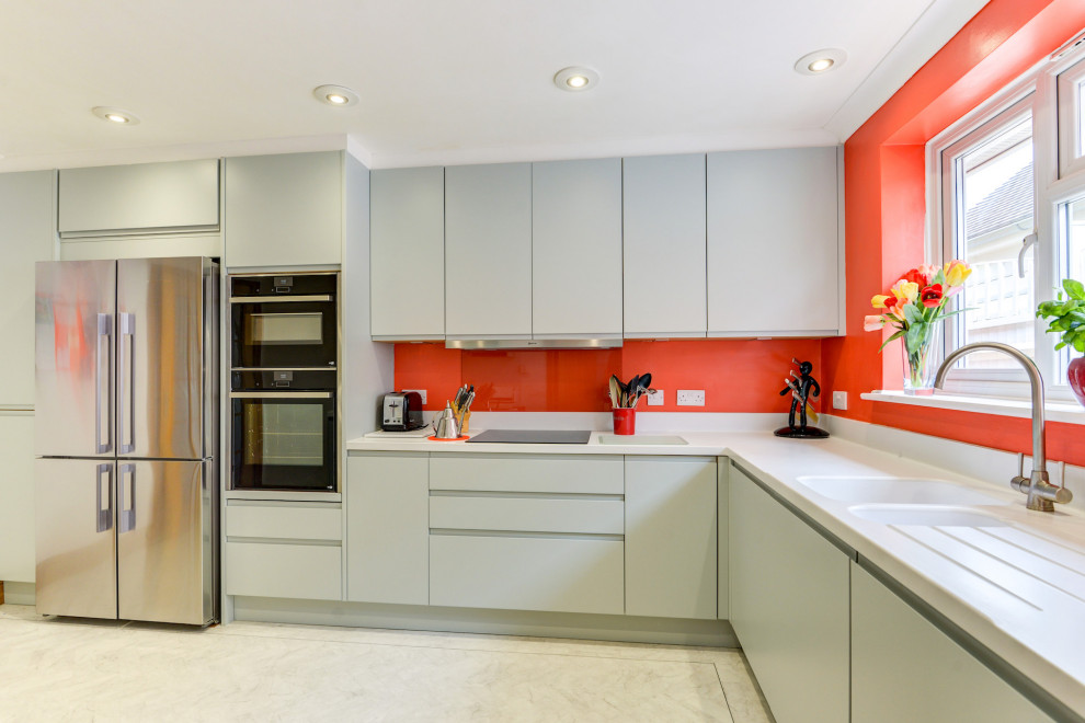 This is an example of a medium sized nautical grey and white l-shaped enclosed kitchen in Sussex with a built-in sink, flat-panel cabinets, green cabinets, composite countertops, orange splashback, glass sheet splashback, black appliances, lino flooring, a breakfast bar, white floors, white worktops and feature lighting.