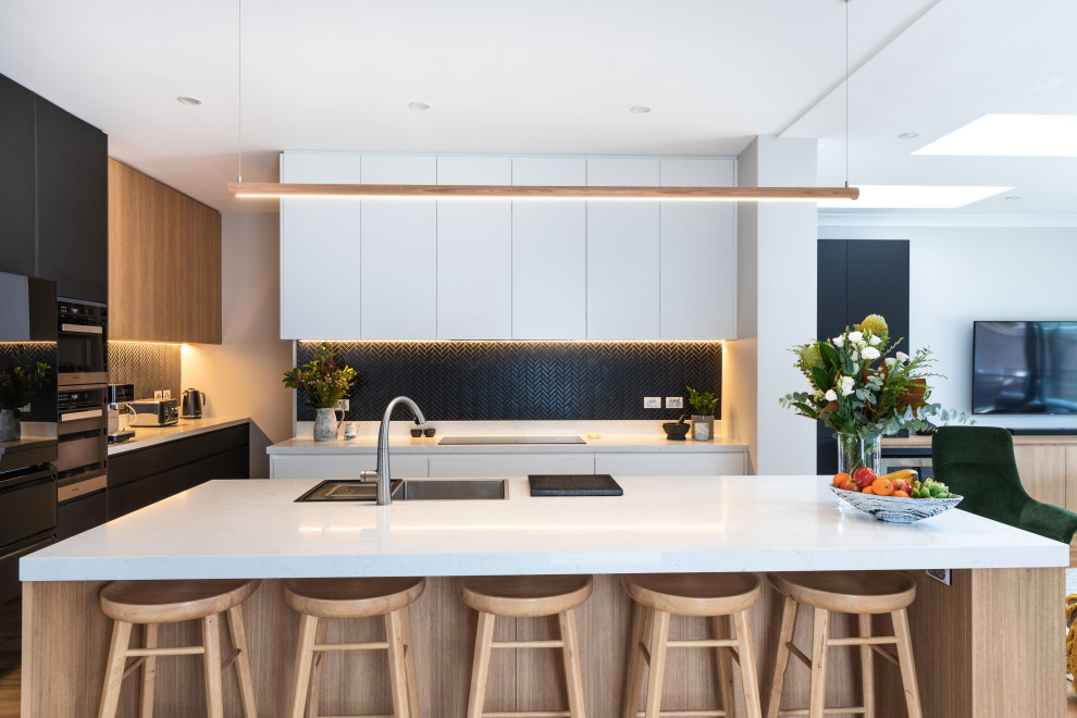 This is an example of a large contemporary kitchen in Sydney with a drop-in sink, flat-panel cabinets, quartz benchtops, black splashback, mosaic tile splashback, black appliances, light hardwood floors, with island and white benchtop.