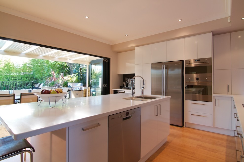 Photo of a mid-sized modern l-shaped eat-in kitchen in Sydney with a triple-bowl sink, white cabinets, glass sheet splashback, stainless steel appliances, with island, flat-panel cabinets, solid surface benchtops, black splashback and light hardwood floors.