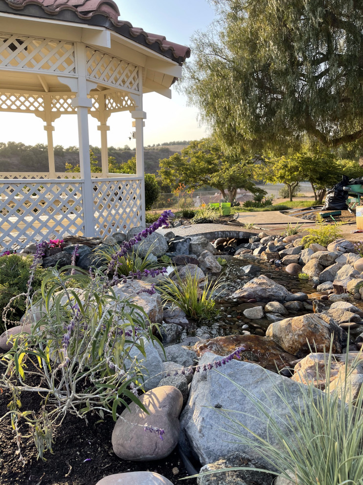 На фото: солнечный, весенний участок и сад среднего размера на заднем дворе в средиземноморском стиле с водопадом, хорошей освещенностью, покрытием из гальки и с деревянным забором с