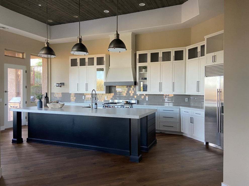 Photo of a large modern single-wall open plan kitchen in Phoenix with a farmhouse sink, shaker cabinets, white cabinets, solid surface benchtops, grey splashback, glass tile splashback, stainless steel appliances, medium hardwood floors, with island, brown floor, white benchtop and timber.