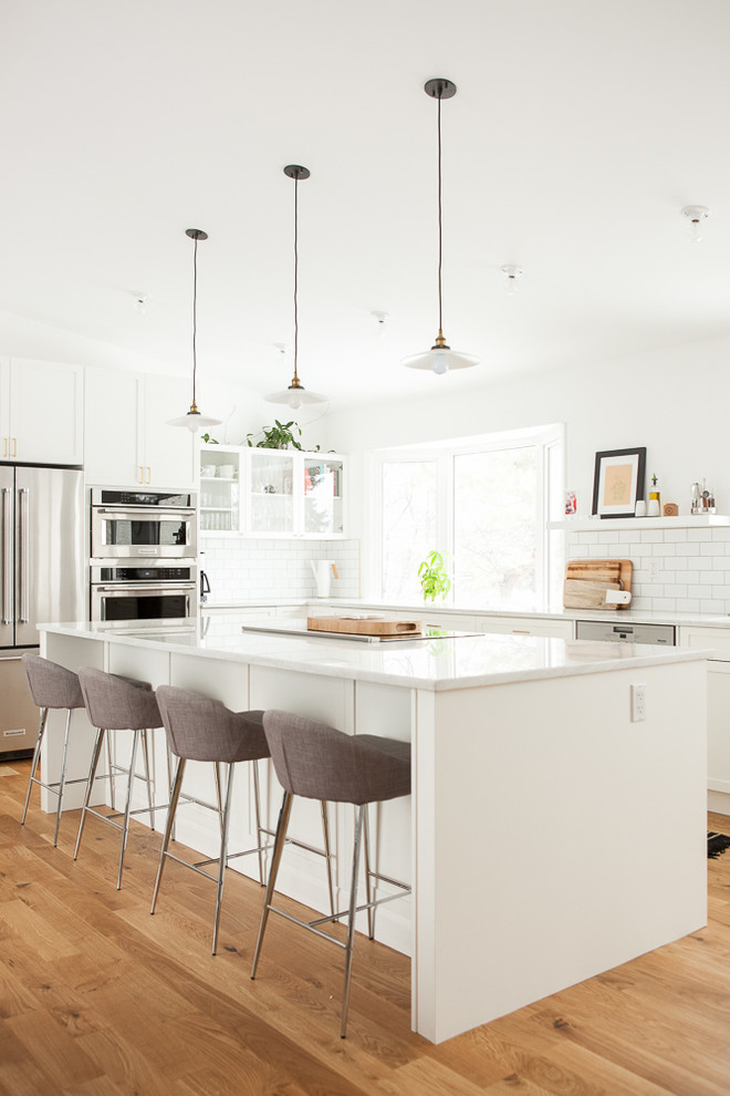 Transitional l-shaped light wood floor kitchen photo with glass-front cabinets, white cabinets, white backsplash, subway tile backsplash, stainless steel appliances and an island
