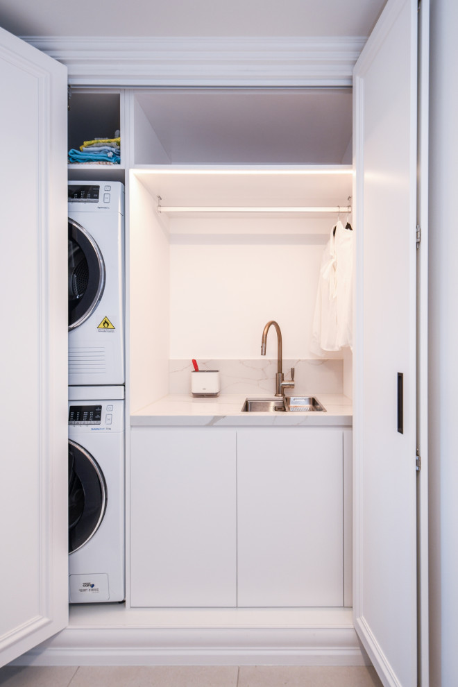 シドニーにあるお手頃価格の小さなトラディショナルスタイルのおしゃれな洗濯室 (I型、アンダーカウンターシンク、フラットパネル扉のキャビネット、白いキャビネット、クオーツストーンカウンター、ベージュキッチンパネル、クオーツストーンのキッチンパネル、白い壁、セラミックタイルの床、上下配置の洗濯機・乾燥機、グレーの床、ベージュのキッチンカウンター) の写真