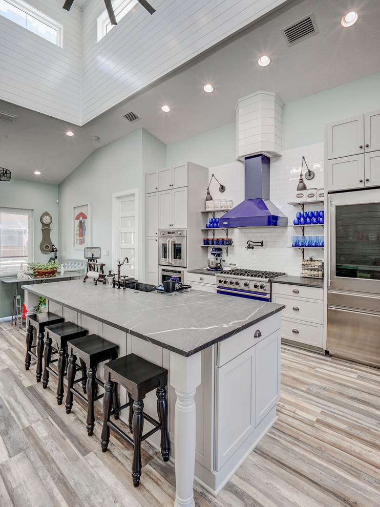 Design ideas for a mid-sized beach style galley open plan kitchen in Tampa with a farmhouse sink, grey cabinets, quartzite benchtops, white splashback, ceramic splashback, stainless steel appliances, ceramic floors, with island, grey floor, grey benchtop and shaker cabinets.