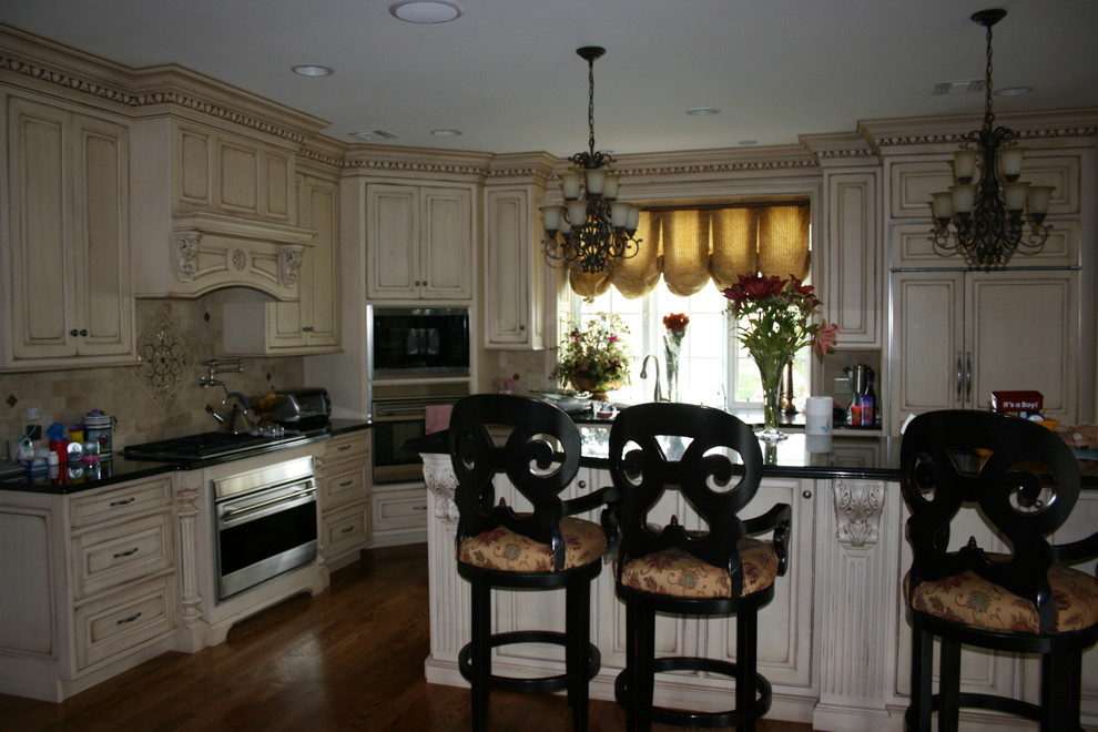 This is an example of a large traditional l-shaped separate kitchen in Philadelphia with an undermount sink, raised-panel cabinets, distressed cabinets, granite benchtops, beige splashback, travertine splashback, stainless steel appliances, dark hardwood floors, with island, brown floor and black benchtop.