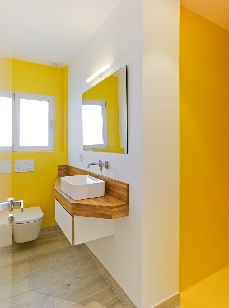 Inspiration for a contemporary bathroom in Other with a wall-mount toilet, yellow walls, a vessel sink, flat-panel cabinets, white cabinets, light hardwood floors and wood benchtops.
