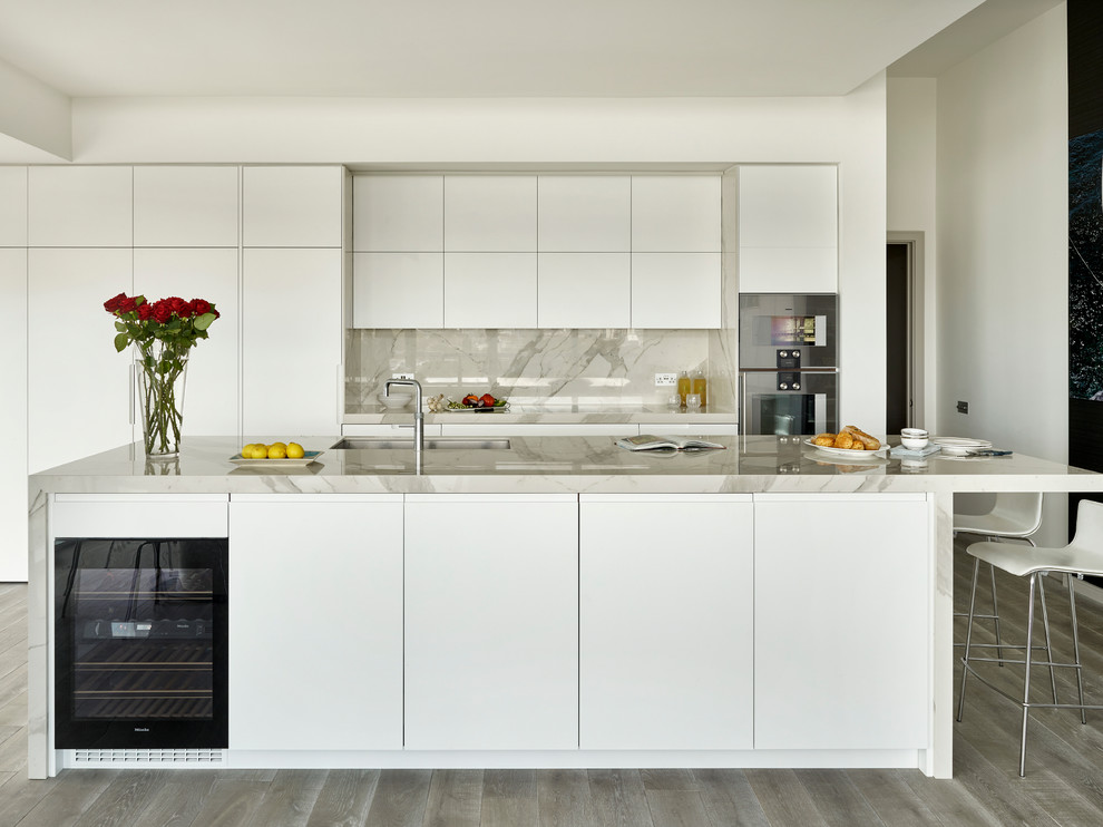 Mid-sized modern single-wall open plan kitchen in London with a double-bowl sink, flat-panel cabinets, white cabinets, marble benchtops, white splashback, stone slab splashback, stainless steel appliances and with island.