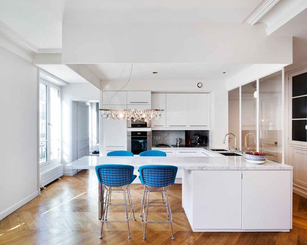 This is an example of a large contemporary u-shaped open plan kitchen in Paris with an undermount sink, flat-panel cabinets, white cabinets, marble benchtops, stainless steel appliances, light hardwood floors and a peninsula.
