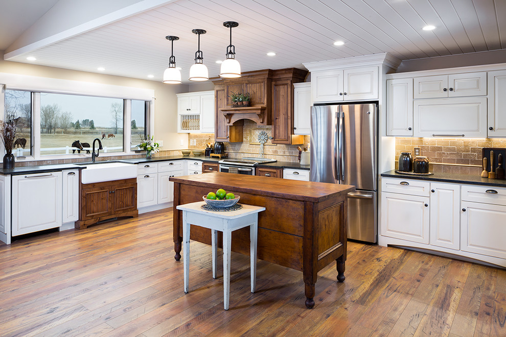 Inspiration for a large farmhouse l-shaped medium tone wood floor kitchen remodel with a farmhouse sink, white cabinets, granite countertops, travertine backsplash, stainless steel appliances, an island, black countertops, raised-panel cabinets and beige backsplash