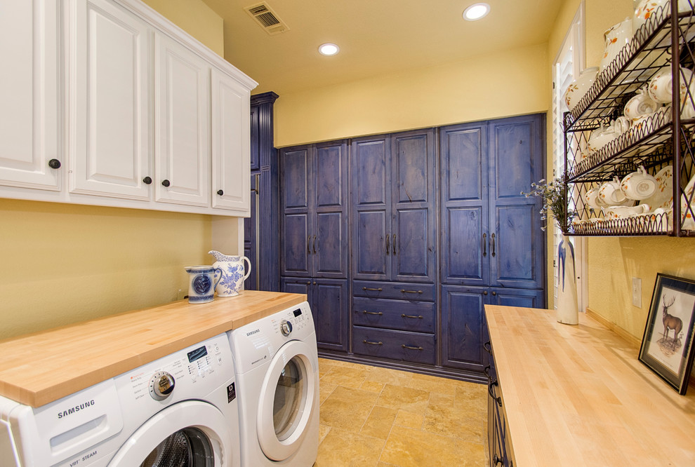 Large country u-shaped dedicated laundry room in Dallas with raised-panel cabinets, white cabinets, wood benchtops, porcelain floors, a side-by-side washer and dryer, brown floor, a drop-in sink, yellow walls and beige benchtop.
