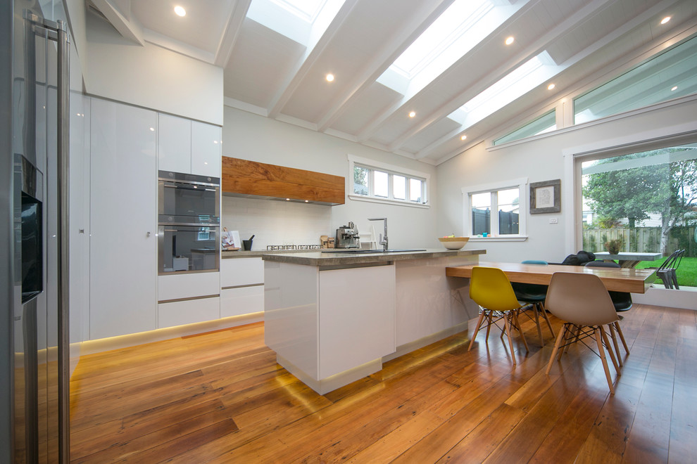 Photo of a large contemporary l-shaped eat-in kitchen in Auckland with white cabinets, concrete benchtops, white splashback, cement tile splashback, stainless steel appliances and with island.