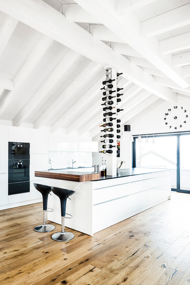 Photo of a large contemporary open plan kitchen in Stuttgart with flat-panel cabinets, white cabinets, white splashback, with island, light hardwood floors, glass sheet splashback and black appliances.