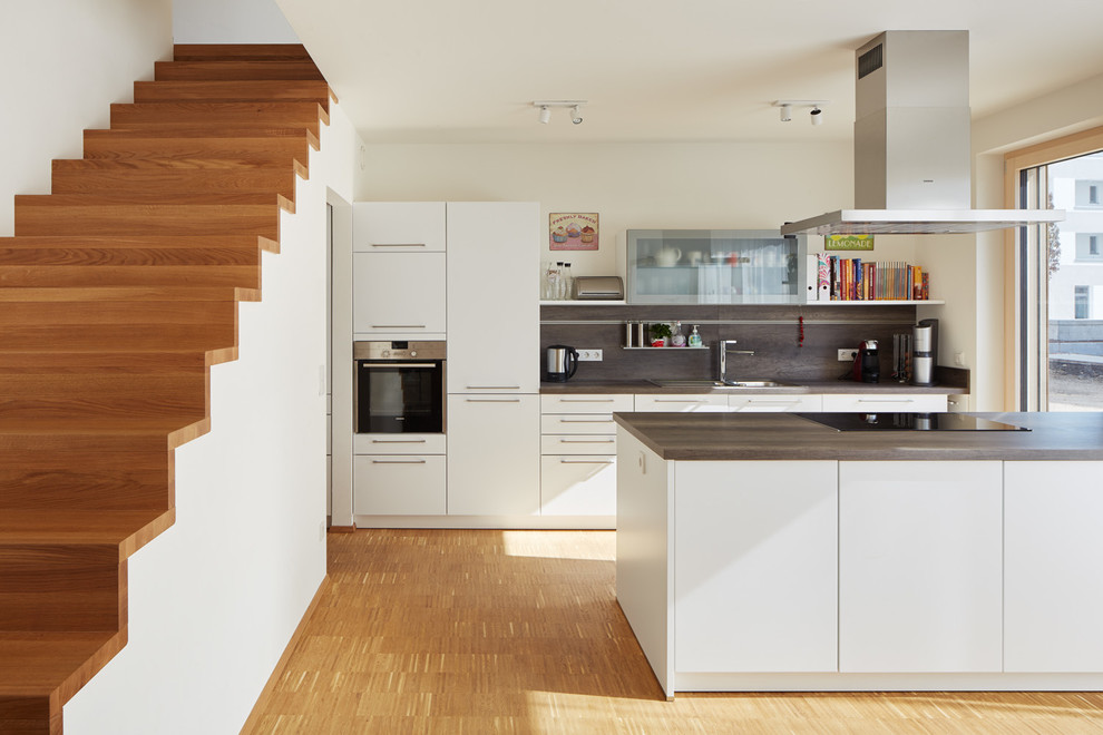 Design ideas for a mid-sized contemporary open plan kitchen in Berlin with a drop-in sink, flat-panel cabinets, white cabinets, wood benchtops, brown splashback, panelled appliances, with island and light hardwood floors.