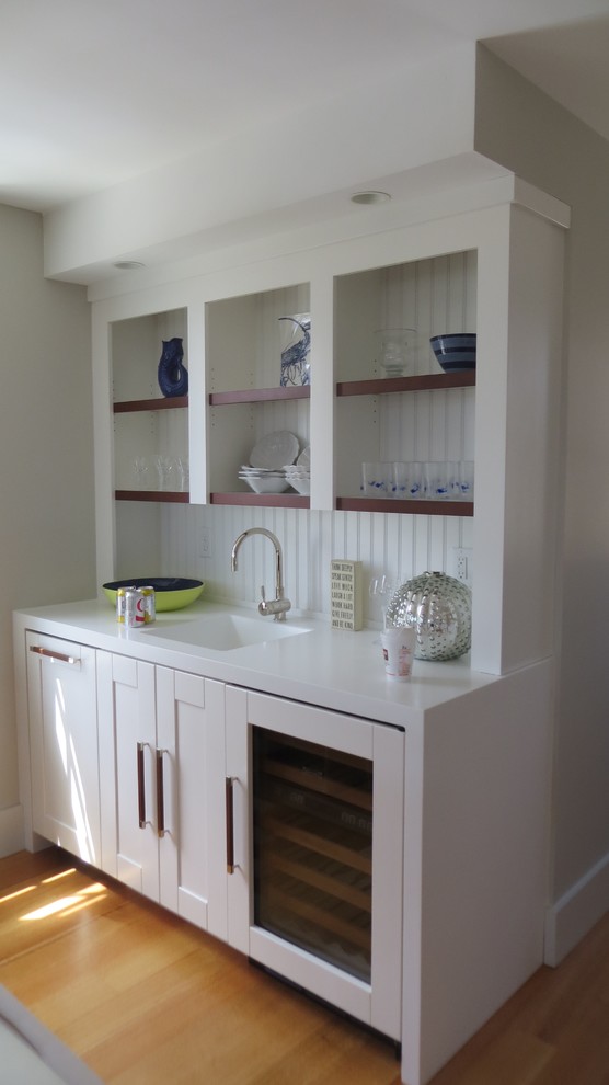 Design ideas for a small beach style single-wall wet bar in Portland Maine with an integrated sink, shaker cabinets, white cabinets, solid surface benchtops, white splashback, timber splashback, light hardwood floors and white benchtop.