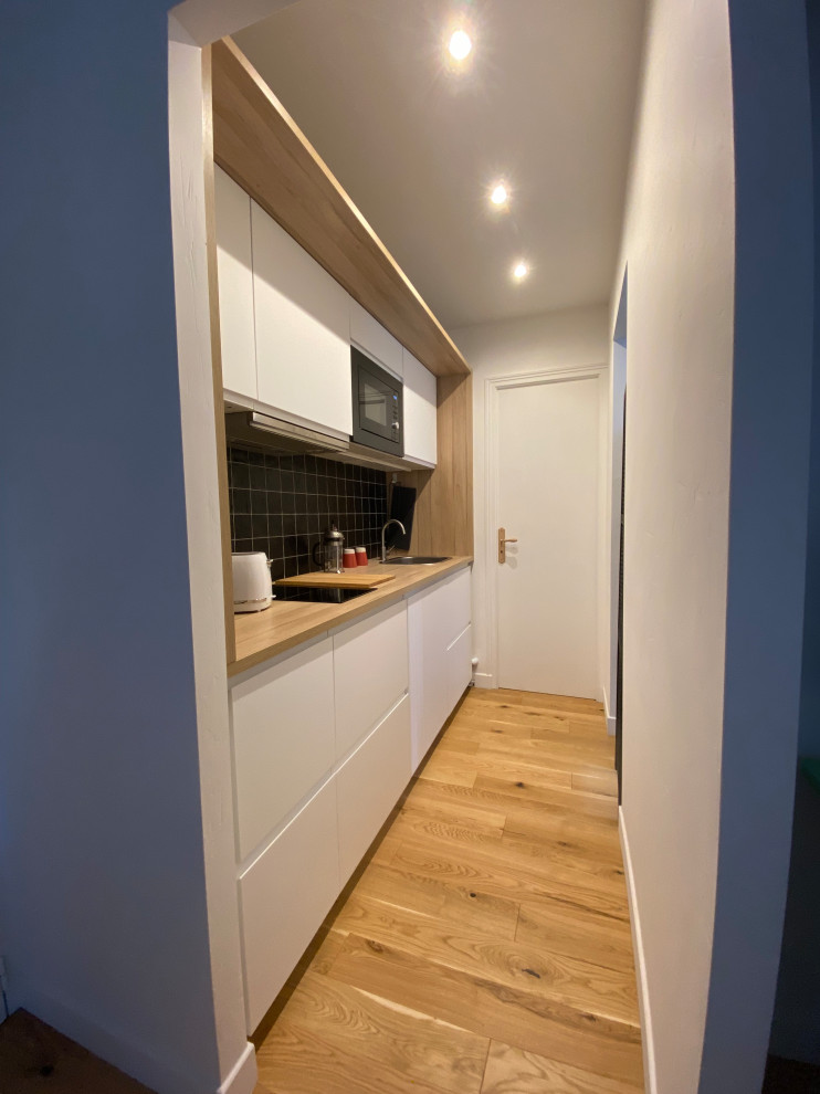 Photo of a small contemporary single-wall kitchen in Other with a single-bowl sink, beaded inset cabinets, white cabinets, laminate benchtops, black splashback, slate splashback and light hardwood floors.
