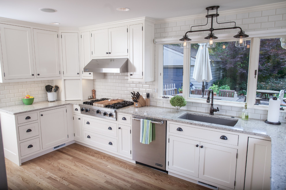 This is an example of a mid-sized industrial l-shaped open plan kitchen in Grand Rapids with an undermount sink, recessed-panel cabinets, white cabinets, quartz benchtops, white splashback, ceramic splashback, stainless steel appliances, light hardwood floors and no island.