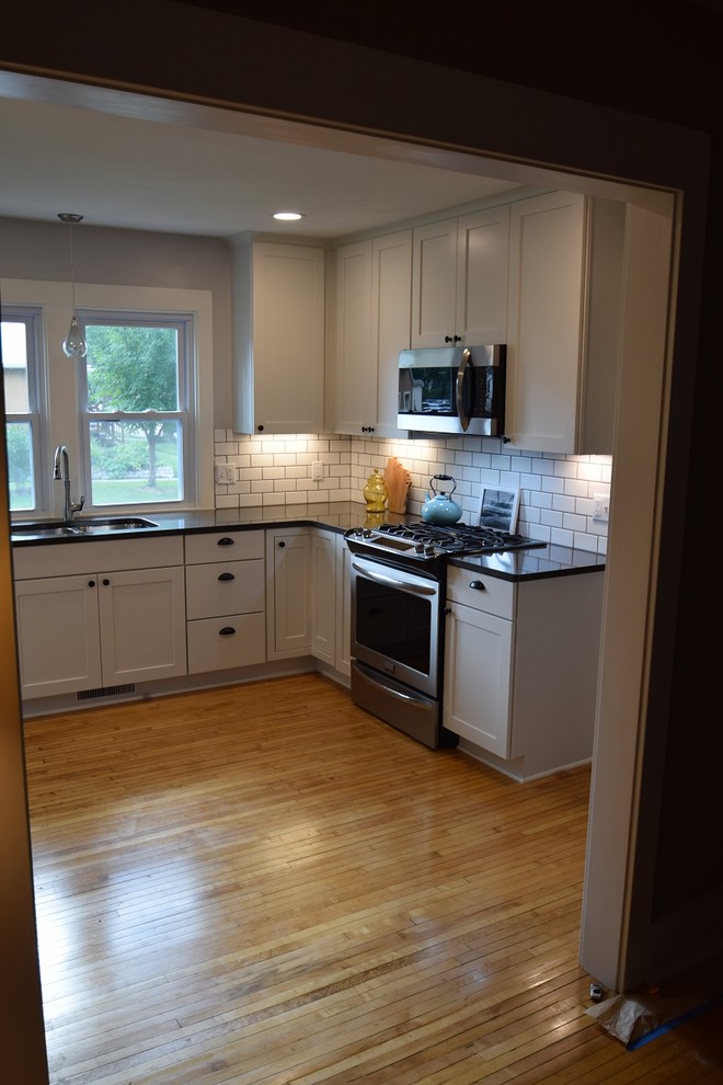 Transitional u-shaped light wood floor kitchen photo with an undermount sink, shaker cabinets, white cabinets, quartz countertops, white backsplash, subway tile backsplash, stainless steel appliances, no island and black countertops