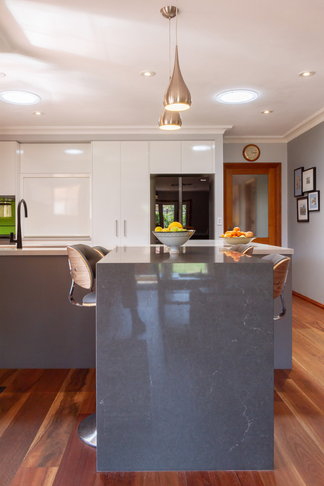This is an example of a mid-sized contemporary u-shaped eat-in kitchen in Melbourne with an undermount sink, flat-panel cabinets, white cabinets, quartz benchtops, green splashback, glass sheet splashback, black appliances, medium hardwood floors, a peninsula, brown floor and white benchtop.