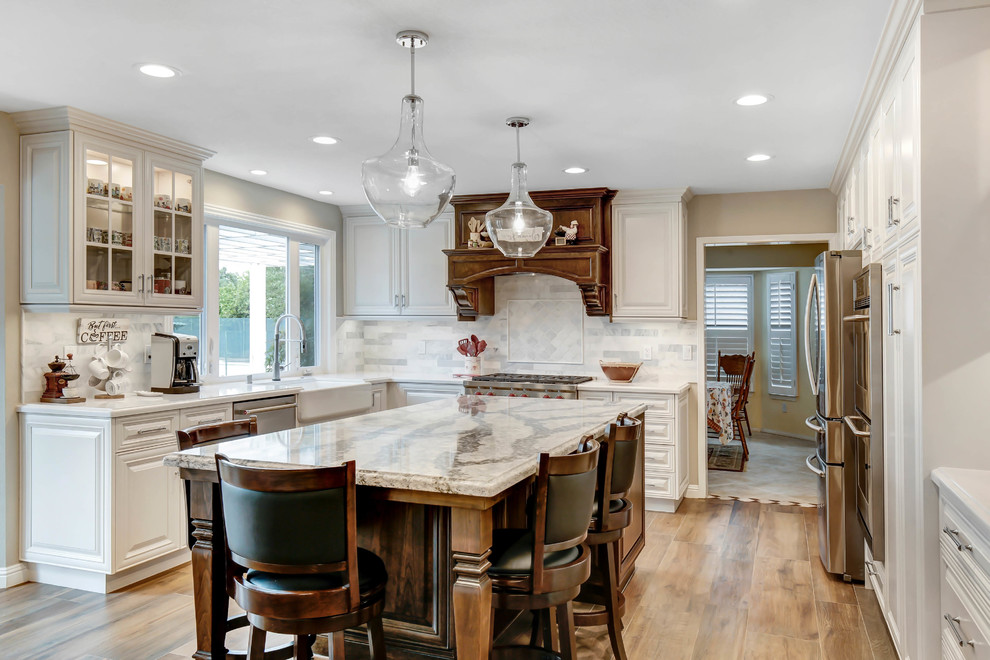 This is an example of a mid-sized traditional u-shaped open plan kitchen in Los Angeles with a farmhouse sink, raised-panel cabinets, white cabinets, quartz benchtops, white splashback, marble splashback, stainless steel appliances, porcelain floors, with island, brown floor and white benchtop.