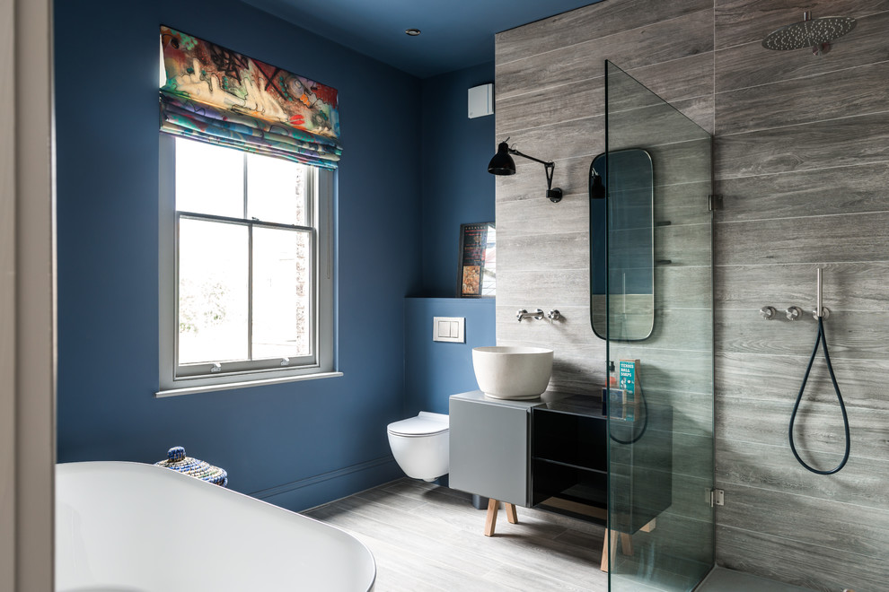 Photo of a mid-sized contemporary master bathroom in London with grey cabinets, a freestanding tub, an open shower, gray tile, stone slab, blue walls, light hardwood floors, a vessel sink, laminate benchtops, a wall-mount toilet, grey floor, an open shower and flat-panel cabinets.
