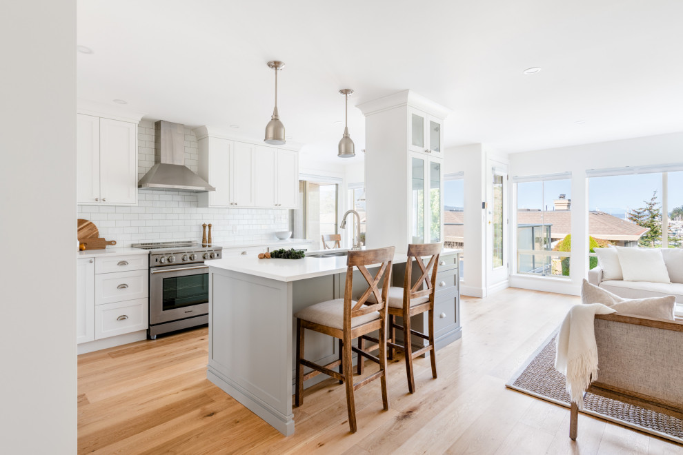 This is an example of a mid-sized country l-shaped open plan kitchen in Vancouver with an undermount sink, shaker cabinets, grey cabinets, quartz benchtops, white splashback, subway tile splashback, stainless steel appliances, medium hardwood floors, with island, brown floor and white benchtop.