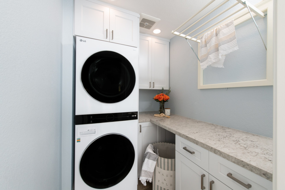 This is an example of a small transitional laundry cupboard in Orange County with recessed-panel cabinets, white cabinets, quartz benchtops, blue walls, vinyl floors, a stacked washer and dryer, brown floor and multi-coloured benchtop.