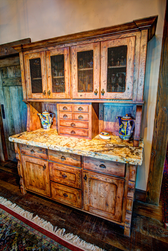 Example of a large mountain style l-shaped dark wood floor eat-in kitchen design with a farmhouse sink, flat-panel cabinets, distressed cabinets, granite countertops, beige backsplash, stone tile backsplash, black appliances and an island