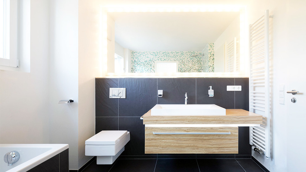 Design ideas for a mid-sized contemporary bathroom in Stuttgart with medium wood cabinets, a drop-in tub, a wall-mount toilet, black tile, stone tile, white walls, slate floors, a vessel sink, wood benchtops and black floor.