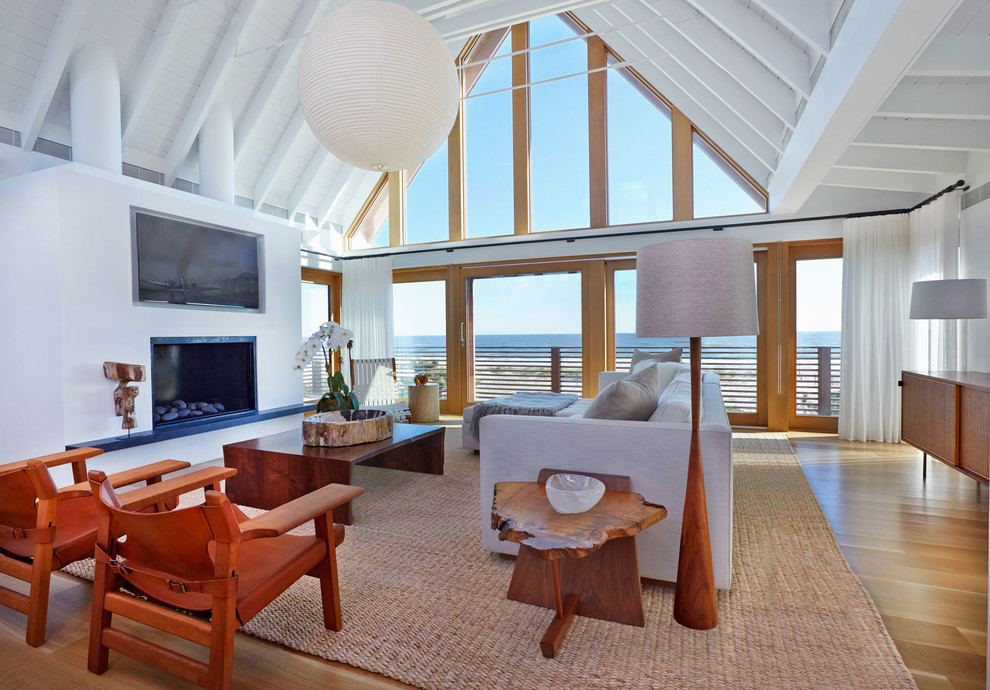 This is an example of a large beach style open concept living room in New York with white walls, medium hardwood floors, a standard fireplace, a concealed tv, a plaster fireplace surround and brown floor.