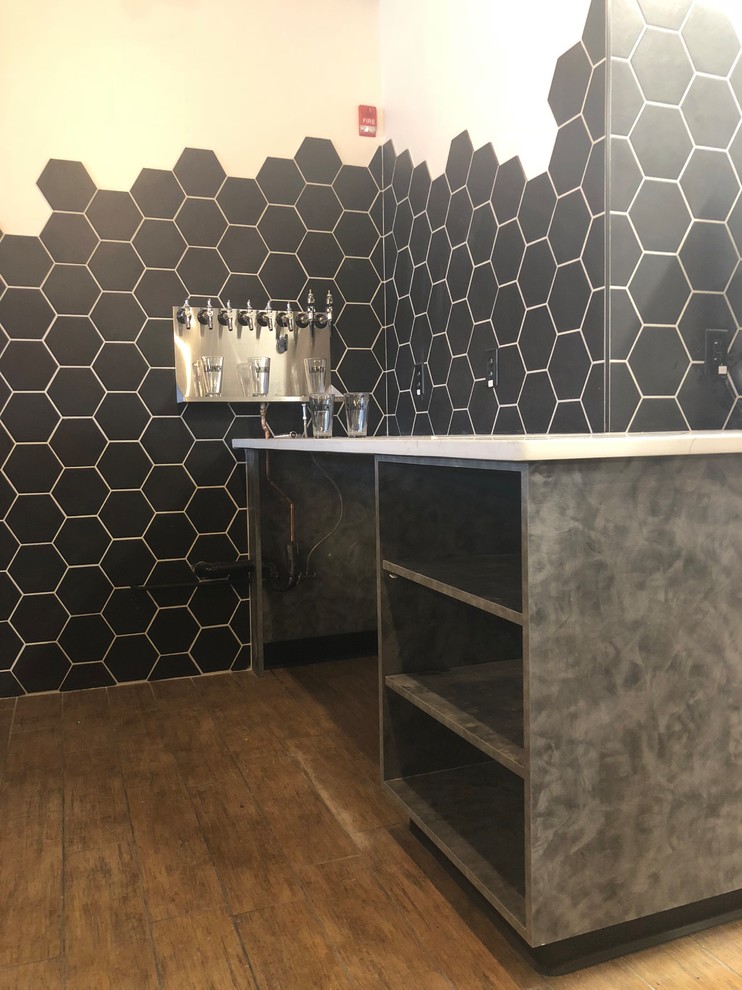 Large industrial l-shaped seated home bar in Omaha with a drop-in sink, open cabinets, grey cabinets, onyx benchtops, black splashback, ceramic splashback, ceramic floors, brown floor and white benchtop.