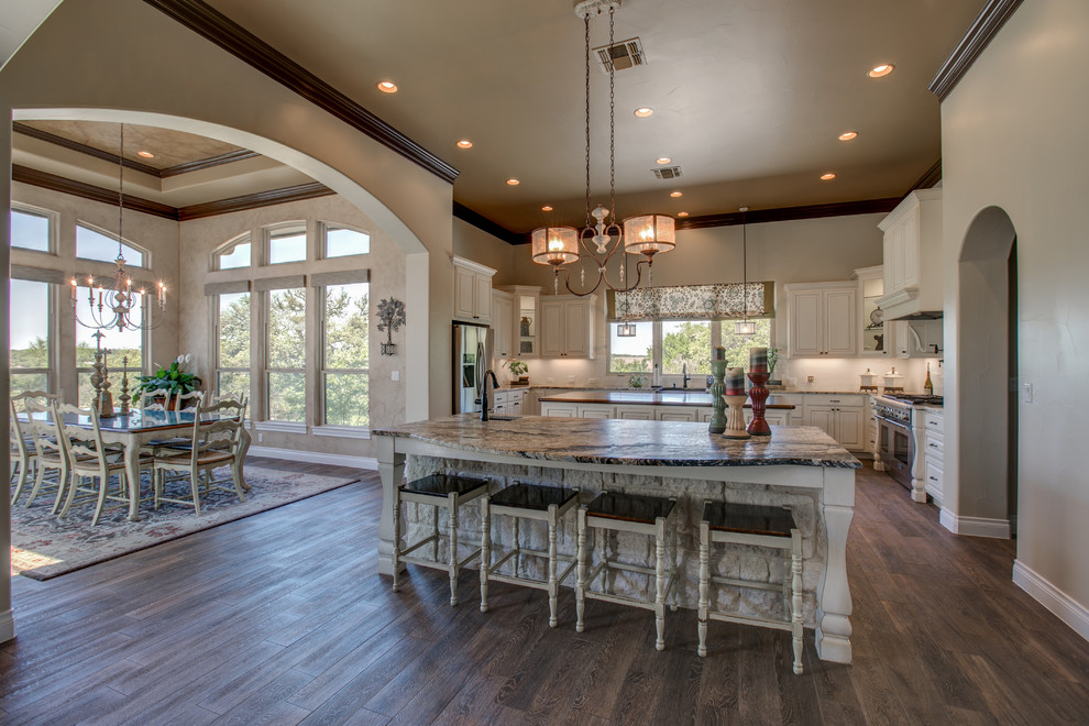 Inspiration for a mid-sized mediterranean u-shaped open plan kitchen in Austin with raised-panel cabinets, white cabinets, granite benchtops, white splashback, ceramic splashback, stainless steel appliances, medium hardwood floors, multiple islands, brown floor and a farmhouse sink.
