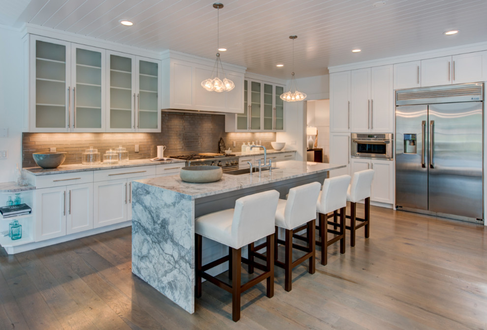 Eat-in kitchen - large transitional l-shaped medium tone wood floor eat-in kitchen idea with a farmhouse sink, glass-front cabinets, white cabinets, gray backsplash, glass tile backsplash, stainless steel appliances, an island and marble countertops