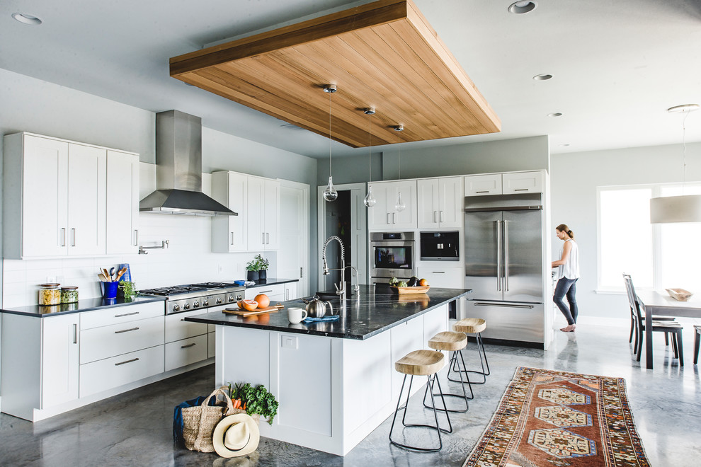 Photo of a transitional l-shaped open plan kitchen in San Francisco with a farmhouse sink, shaker cabinets, white cabinets, white splashback, stainless steel appliances, concrete floors, with island, grey floor and black benchtop.