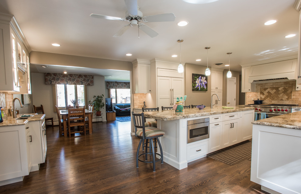 Example of a large transitional u-shaped medium tone wood floor and brown floor eat-in kitchen design with an undermount sink, shaker cabinets, white cabinets, granite countertops, beige backsplash, travertine backsplash, paneled appliances and an island