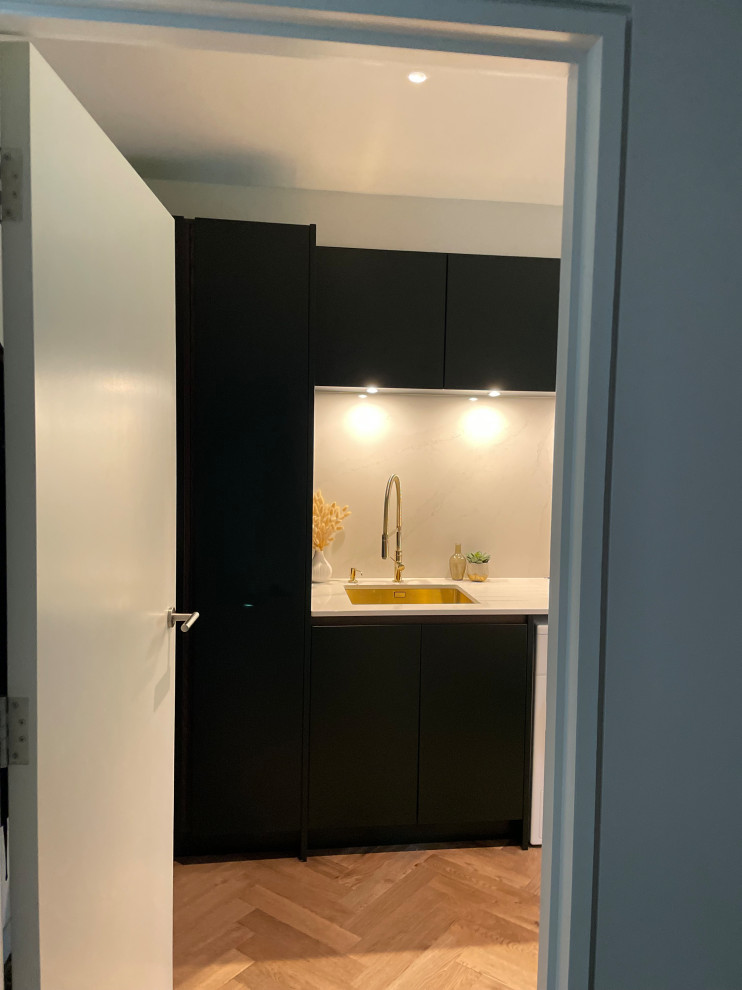 This is an example of a mid-sized contemporary galley dedicated laundry room in Hertfordshire with an integrated sink, flat-panel cabinets, blue cabinets, quartzite benchtops, white splashback, engineered quartz splashback, white walls, medium hardwood floors, a side-by-side washer and dryer, brown floor and white benchtop.