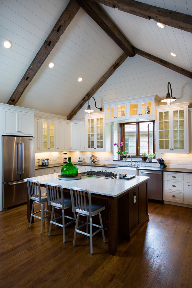 Design ideas for a mid-sized country l-shaped open plan kitchen in Atlanta with glass-front cabinets, white cabinets, stainless steel appliances, white splashback, subway tile splashback, dark hardwood floors, with island, a farmhouse sink, marble benchtops and brown floor.