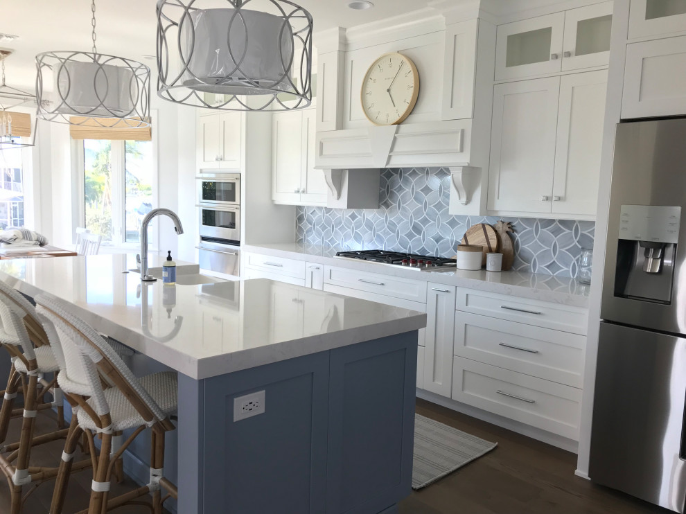 This is an example of a mid-sized beach style kitchen with a farmhouse sink, shaker cabinets, blue cabinets, quartz benchtops, blue splashback, glass tile splashback, stainless steel appliances, medium hardwood floors, with island, brown floor and white benchtop.