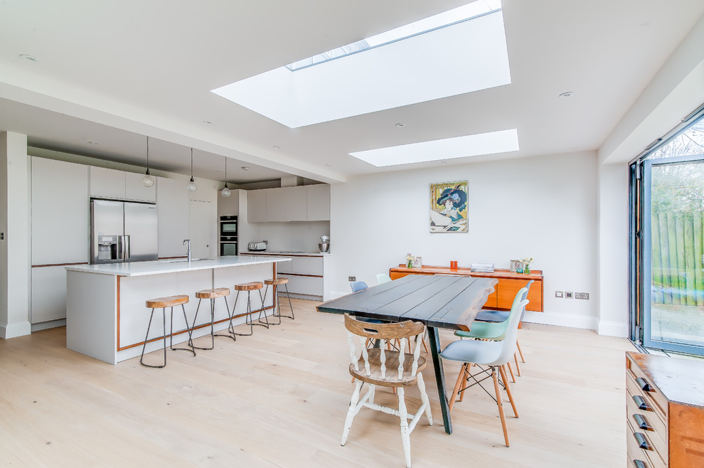 Mid-sized contemporary l-shaped eat-in kitchen in London with an undermount sink, flat-panel cabinets, white cabinets, solid surface benchtops, white splashback, stainless steel appliances, with island, beige floor, light hardwood floors and white benchtop.