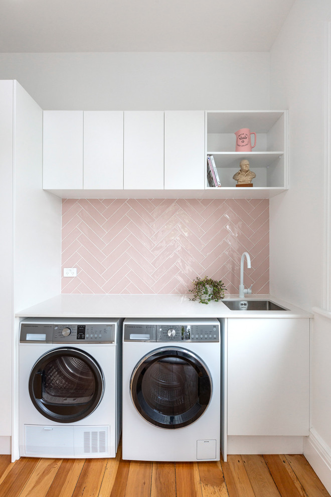 Mid-sized modern single-wall dedicated laundry room in Auckland with an undermount sink, flat-panel cabinets, white cabinets, light hardwood floors, pink splashback, subway tile splashback, a side-by-side washer and dryer and white benchtop.
