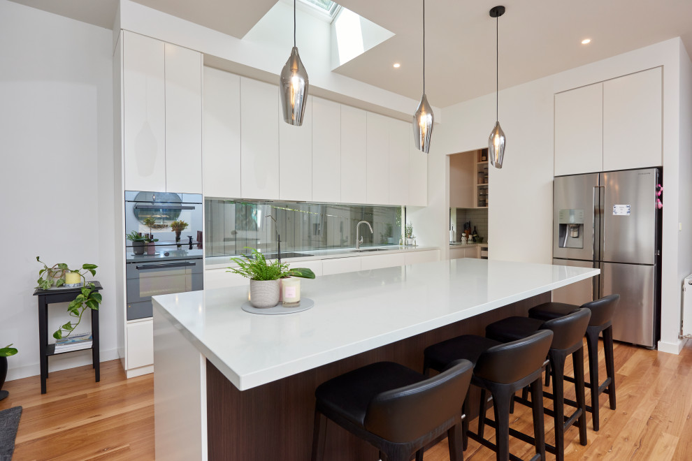 Inspiration for a contemporary l-shaped kitchen in Melbourne with an undermount sink, flat-panel cabinets, white cabinets, window splashback, stainless steel appliances, light hardwood floors, with island, beige floor and white benchtop.