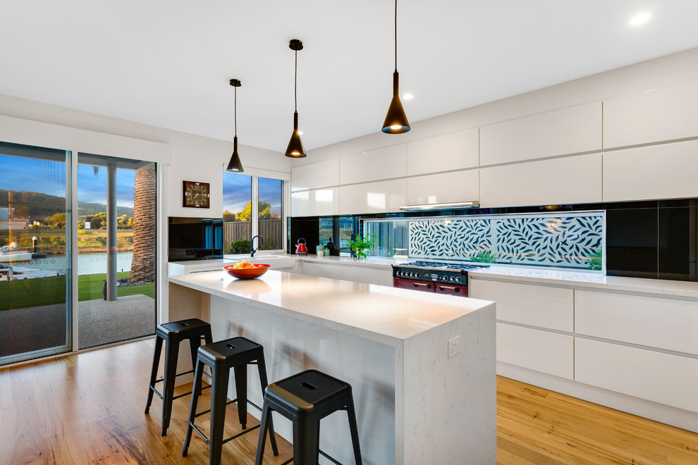 Large contemporary l-shaped open plan kitchen in Melbourne with a farmhouse sink, flat-panel cabinets, white cabinets, window splashback, coloured appliances, light hardwood floors, with island, beige floor, quartz benchtops, black splashback and grey benchtop.