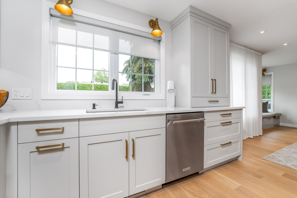 This is an example of a large contemporary l-shaped open plan kitchen in Toronto with a double-bowl sink, shaker cabinets, grey cabinets, quartz benchtops, white splashback, porcelain splashback, stainless steel appliances, light hardwood floors, with island, brown floor and white benchtop.