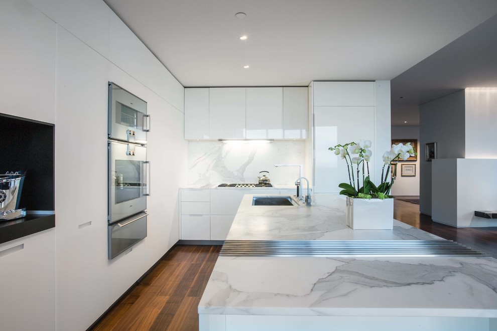 Design ideas for a large modern l-shaped open plan kitchen in Salt Lake City with an undermount sink, flat-panel cabinets, white cabinets, marble benchtops, white splashback, marble splashback, panelled appliances, medium hardwood floors, with island and brown floor.