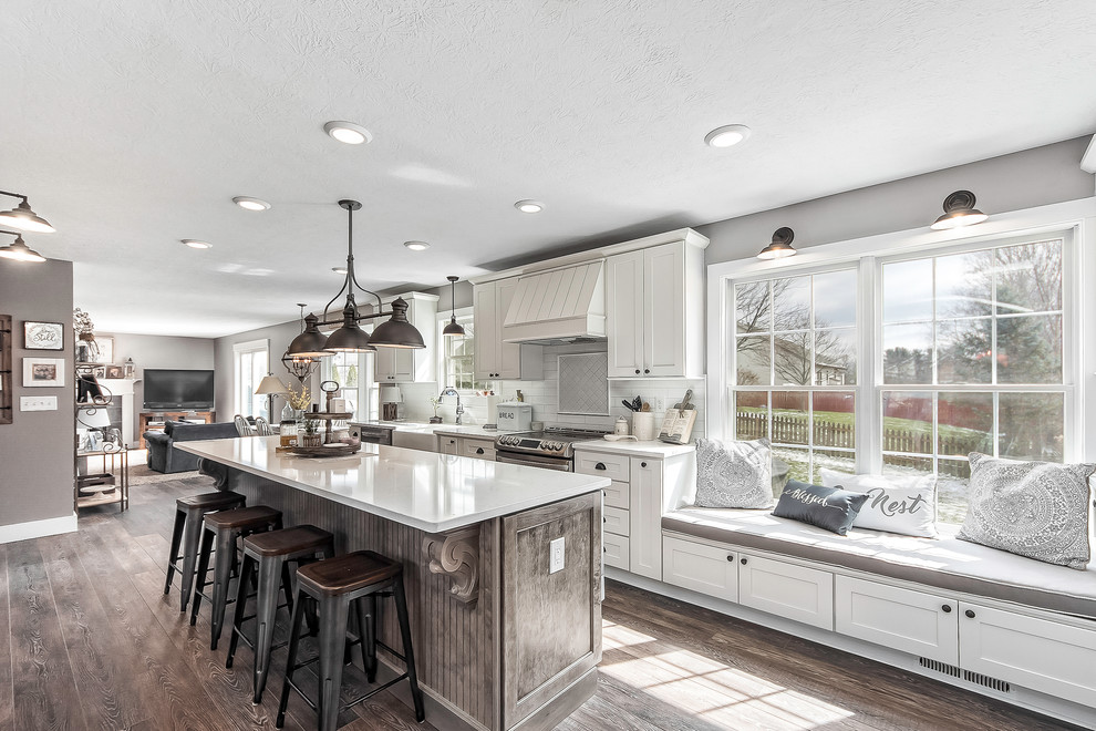 Photo of a large country open plan kitchen in Columbus with a farmhouse sink, shaker cabinets, white cabinets, quartz benchtops, white splashback, subway tile splashback, stainless steel appliances, medium hardwood floors, with island, brown floor and white benchtop.