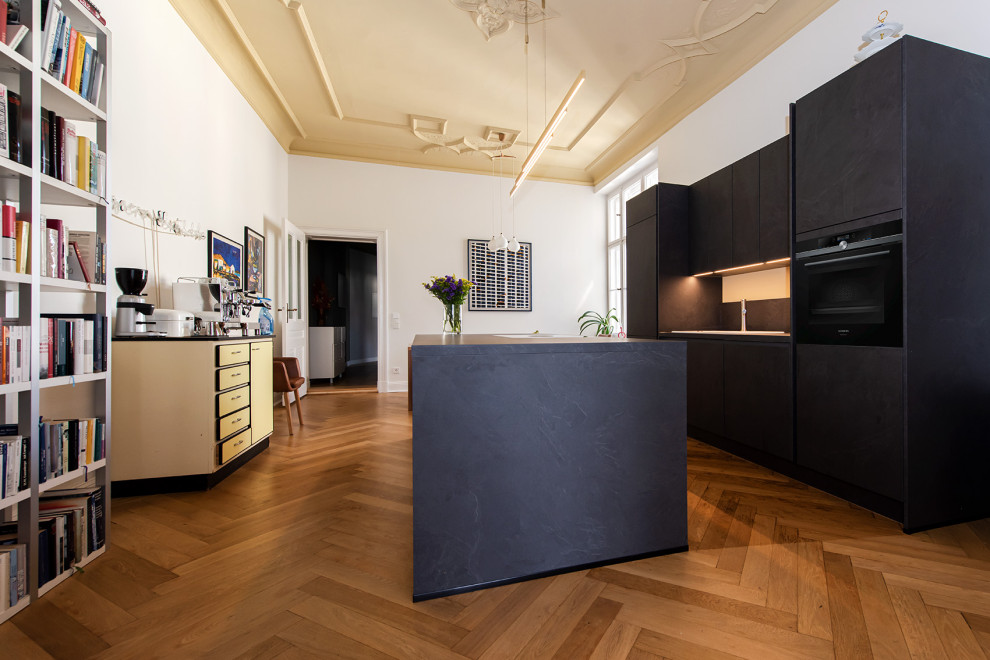 Large eclectic single-wall kitchen in Berlin with a drop-in sink, grey cabinets, laminate benchtops, grey splashback, black appliances, light hardwood floors, with island, brown floor and grey benchtop.