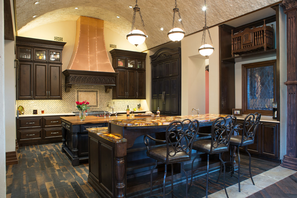 Large transitional l-shaped eat-in kitchen in Austin with a farmhouse sink, raised-panel cabinets, dark wood cabinets, onyx benchtops, multi-coloured splashback, cement tile splashback, panelled appliances, dark hardwood floors, multiple islands, brown floor and brown benchtop.