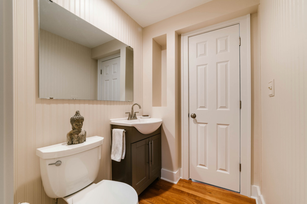 Photo of a small arts and crafts powder room in Chicago with shaker cabinets, brown cabinets, a one-piece toilet, beige walls, medium hardwood floors, a trough sink, solid surface benchtops, brown floor and white benchtops.