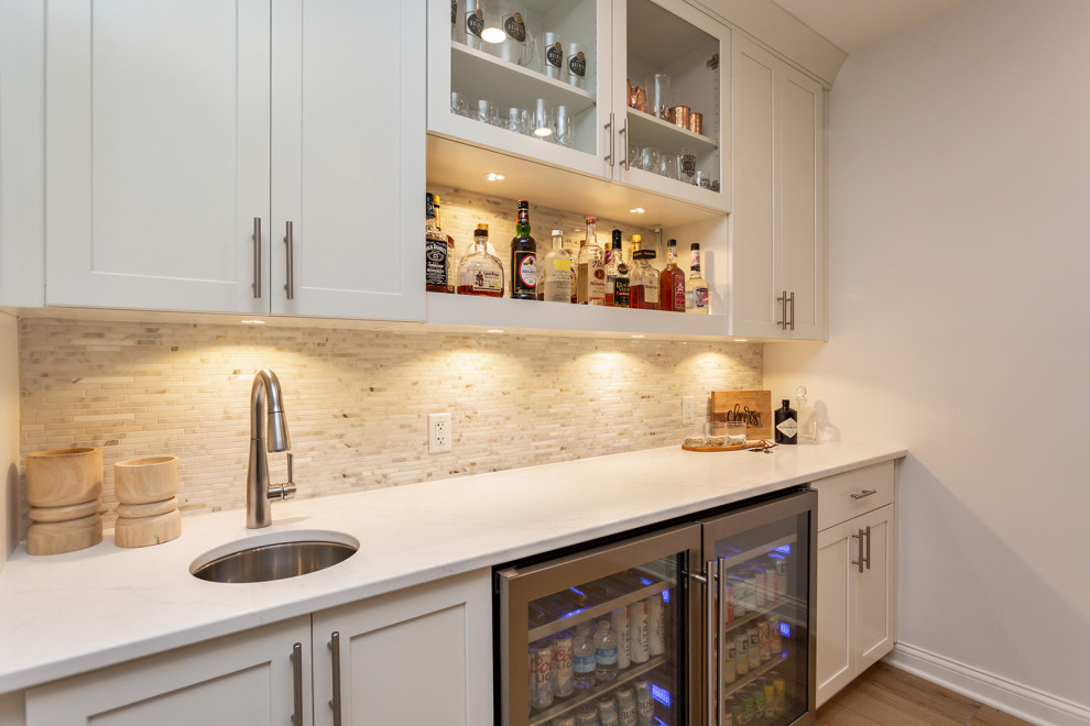 Design ideas for a large classic single-wall wet bar in Other with a submerged sink, recessed-panel cabinets, white cabinets, engineered stone countertops, white splashback, mosaic tiled splashback, light hardwood flooring, brown floors and white worktops.